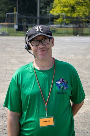 Man in green committee t-shirt and headset smiling outdoors at a sporting event on a sunny day.