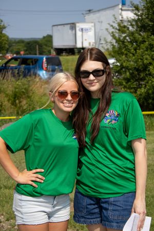 Two young women in matching green t-shirts and sunglasses smile outdoors at a sunny event.