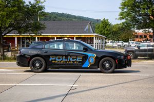 Wellsville NY police car, a black Dodge Charger patrol vehicle parked on a street with green trees in background.