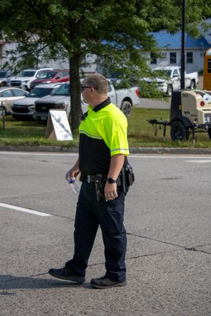 A security officer in a neon yellow vest walks across a parking lot, wearing a duty belt and black pants.
