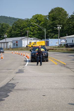 Police officer in reflective vest directs traffic near orange cones on a road with trees and buildings in background.