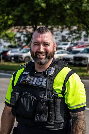 Smiling male police officer in yellow high-visibility vest and tactical gear standing outdoors near a parking lot.