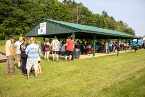 Crowd of people gathered under a large green pavilion shelter on a sunny day at an outdoor park event.