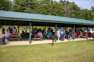 Large group gathered under a green-roofed outdoor pavilion with red picnic tables on a sunny day.