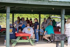 Group of people gathered under a green pavilion with red picnic tables at an outdoor community event.