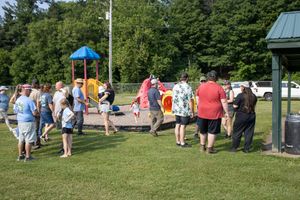 Group of people gathering at a park playground on a sunny day with green trees in the background.