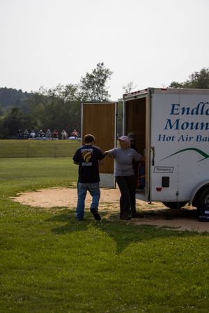 Two people unloading equipment from an Endless Mountains Hot Air Balloon trailer on a grassy field.