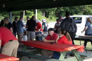 Group of people gathered at outdoor picnic tables under a pavilion for a casual company cookout event.