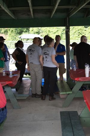 Group of people socializing under a covered outdoor pavilion with red and green picnic tables at a casual gathering.