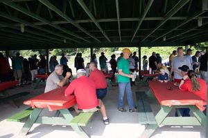 People gather under a covered outdoor pavilion with red and green picnic tables at a casual group event.