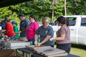 Volunteers serving food outdoors at a community event, working together at tables under shade trees.