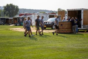 Group of people walking on grass at outdoor festival with food trucks and trailers on a sunny day.