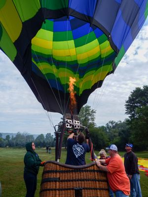 People inflate a colorful green and blue striped hot air balloon with a propane burner flame on a grassy field.