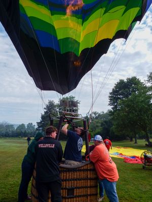 Crew members inflate a colorful blue and green hot air balloon on a grassy field, with flame visible overhead.