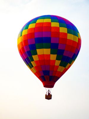 Colorful rainbow checkered hot air balloon floating in a clear sky with passengers in the wicker basket below.