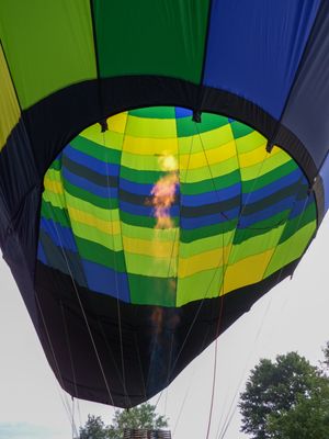 Colorful hot air balloon interior with green, blue, and yellow stripes illuminated by a bright burner flame against a cloudy sky.