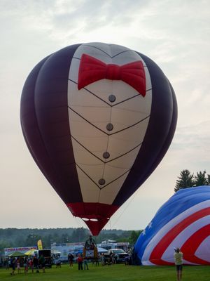 A novelty hot air balloon designed to look like a tuxedo with a red bow tie floats at an outdoor balloon festival.