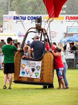 Hot air balloon basket with Thank You banner at a fair, surrounded by people, with Snow Cones and Popcorn signs in background.