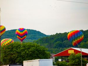 Colorful hot air balloons float above treetops near a red barn with green hills in the background.