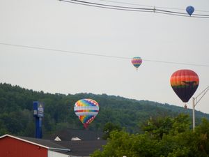 Four colorful hot air balloons float above red buildings and green hills on a cloudy day at a balloon festival.