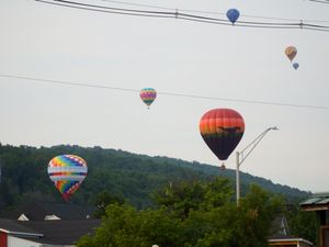 Multiple colorful hot air balloons float over a small town with tree-covered hills in the background during a balloon festival.