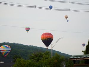 Multiple colorful hot air balloons floating over a tree-covered hillside near a small town on a cloudy day.