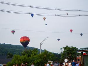 Colorful hot air balloons fill the sky above a small town during a summer balloon festival, with spectators watching below.