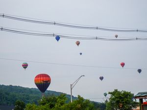 Colorful hot air balloons float over a small town with power lines and green trees under a cloudy sky.
