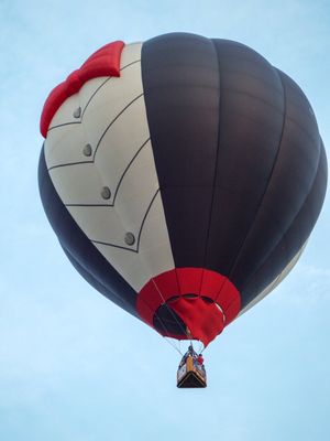 Black, white, and red hot air balloon with decorative design soaring against a clear blue sky with passengers in basket.
