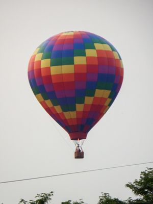 Colorful checkered hot air balloon with rainbow pattern flying over treetops against a cloudy sky.