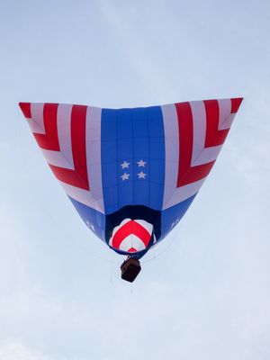 Patriotic red, white, and blue American flag hot air balloon soaring against a cloudy sky, viewed from below.