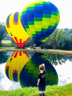 A young boy stands before two vibrant hot air balloons reflecting on a calm pond surrounded by lush green trees.