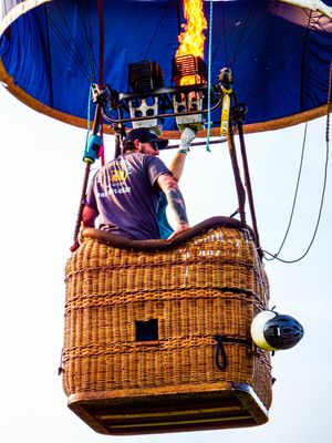 Tattooed pilot firing burner in wicker basket of colorful hot air balloon against bright sky.