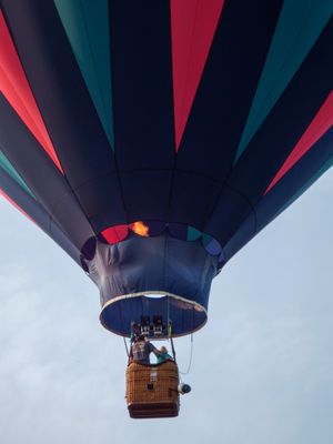 A colorful hot air balloon with red, teal, and navy stripes soars against a cloudy sky, viewed from below.