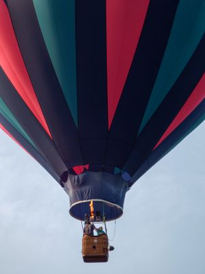 A colorful striped hot air balloon with red, teal, and dark panels soars against a cloudy sky with passengers in basket.