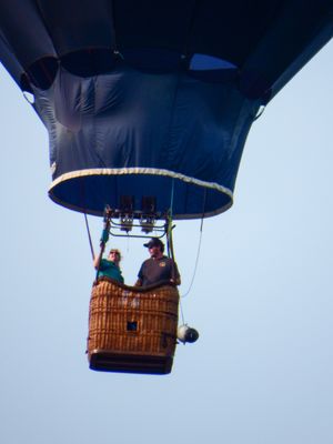 Two people ride in a wicker basket beneath a dark blue hot air balloon floating against a clear sky.