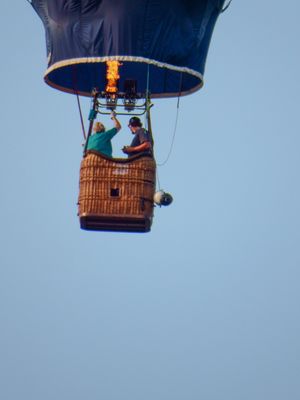 Two people ride in a wicker basket gondola of a blue hot air balloon with a visible flame burner against a clear sky.