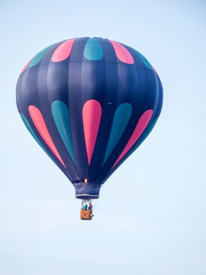 A colorful hot air balloon with pink and teal teardrop patterns floats against a clear blue sky with passengers in basket.