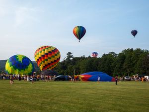 Colorful hot air balloons launch and float over a green field at a summer balloon festival with crowds of spectators.