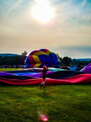 Woman stands among colorful hot air balloons being inflated on green grass at a sunny outdoor balloon festival.
