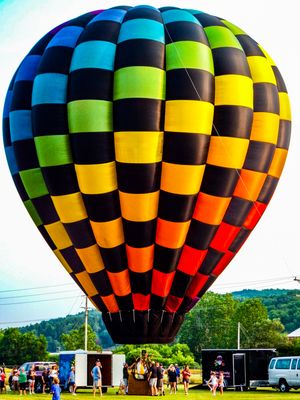 Colorful checkered hot air balloon inflated at an outdoor festival with spectators and vehicles on green grass.