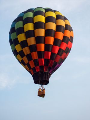 Colorful checkered hot air balloon with rainbow pattern floating against a blue sky with passengers in the basket.
