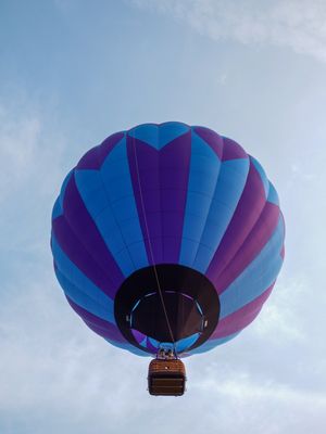 Purple and blue hot air balloon with wicker basket soaring against a partly cloudy sky.