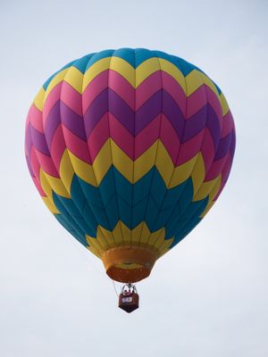 Colorful hot air balloon with chevron pattern in pink, yellow, purple, and teal soaring against a pale sky.