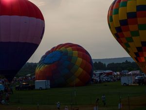 Colorful hot air balloons being inflated at a twilight balloon festival on a green field with spectators.