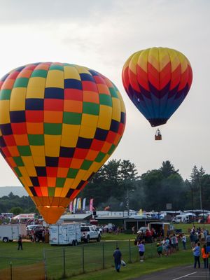 Two colorful hot air balloons at a festival, one checkered yellow and blue, another red and orange, floating above a crowd.