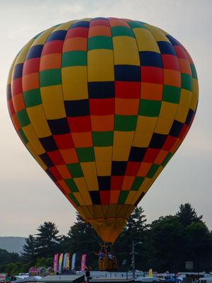 Colorful checkered hot air balloon in yellow, red, green, and blue rising above treetops at an outdoor festival.
