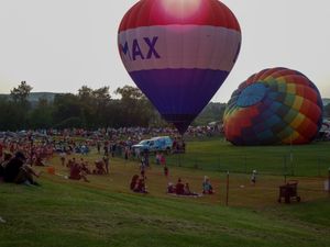 Two colorful hot air balloons, including a RE/MAX branded one, inflate at a crowded outdoor festival on a grassy field at dusk.