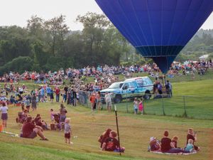 Large crowd gathers at outdoor hot air balloon festival with blue balloon launching over green hillside.