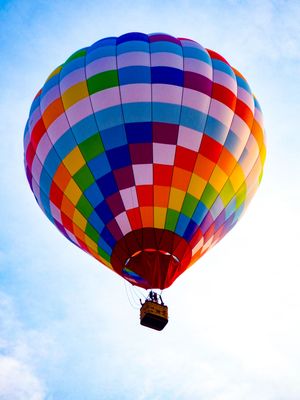 Colorful rainbow checkered hot air balloon soaring against a blue sky with passengers in the wicker basket below.